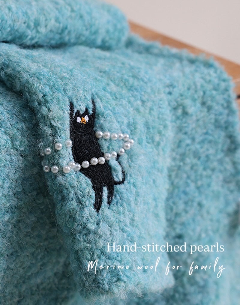 Blue fabric with a black cat embroidered with hand-stitched pearls and text 'Hand-stitched pearls Merino wool for family'.