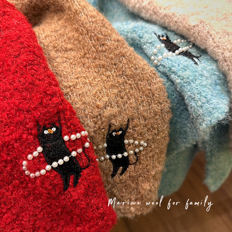 Stack of colorful knitted fabrics with embroidered black cats holding pearls, labeled 'Merino wool for family'.